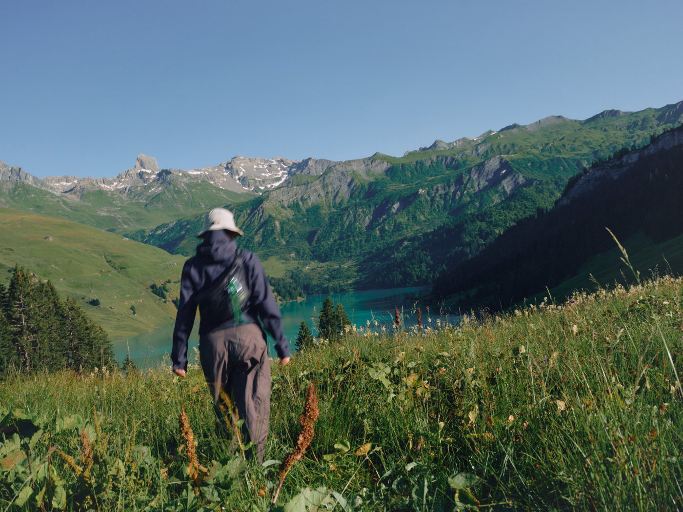 A walker in a wide-brimmed hat crosses a high alpine meadow toward a turquoise lake